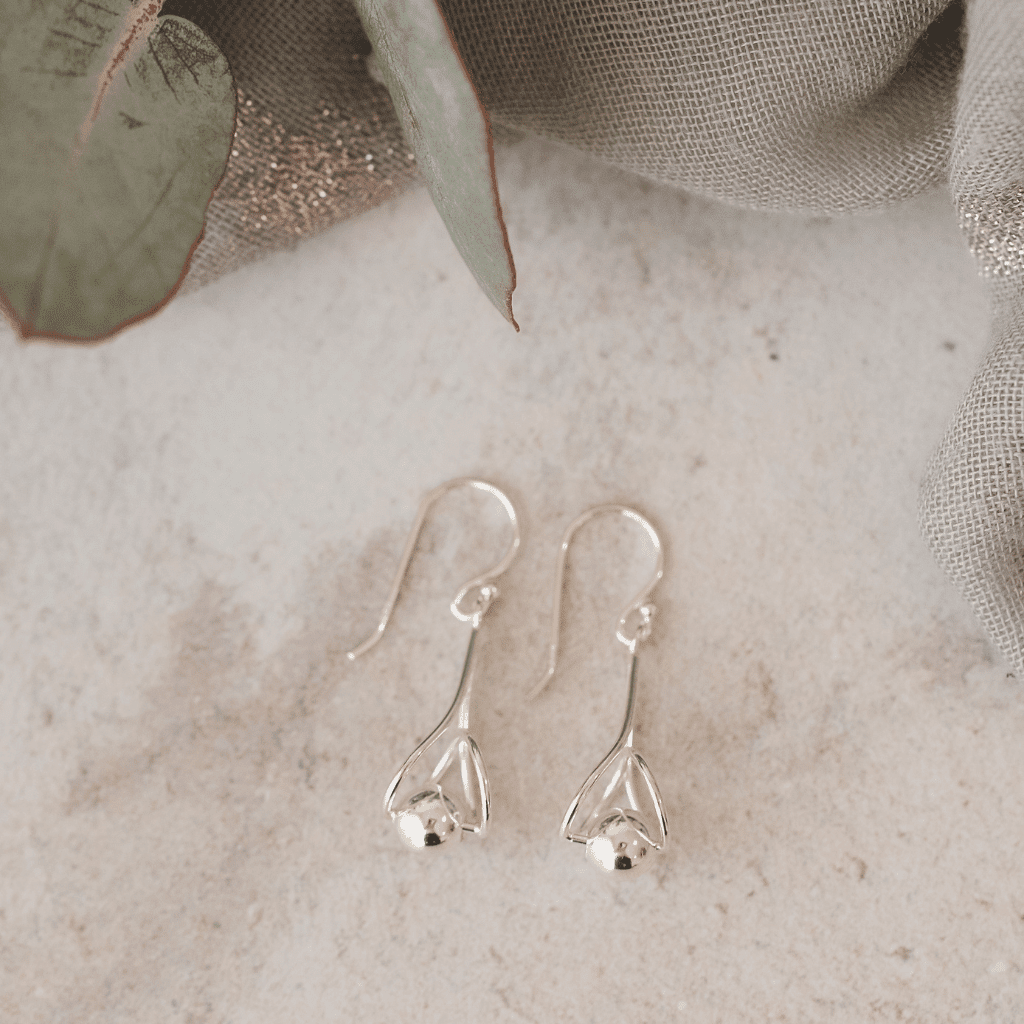 A pair of silver hook earrings, each featuring a twisted, tapering stem holding a polished silver ball. They are displayed on a light gray surface with a soft, green leafy branch in the background.