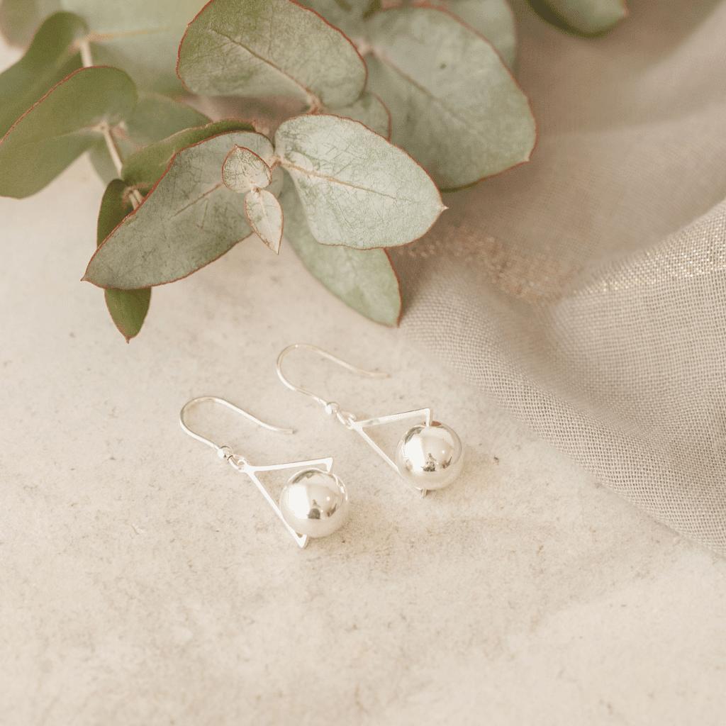 A pair of silver hook earrings, each featuring a twisted, tapering stem holding a polished silver ball. They are displayed on a light gray surface with a soft, green leafy branch in the background.